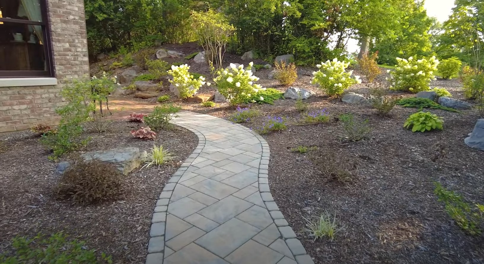 Professional landscape installation featuring natural stone pathways and flowering plants Curved stone pathway through professionally landscaped garden with white flowering shrubs