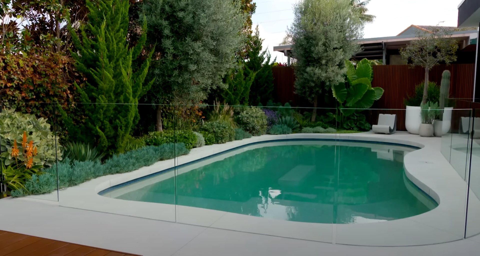 Contemporary backyard pool with professional landscape design and safety features Modern pool area with glass safety fencing surrounded by mature landscaping